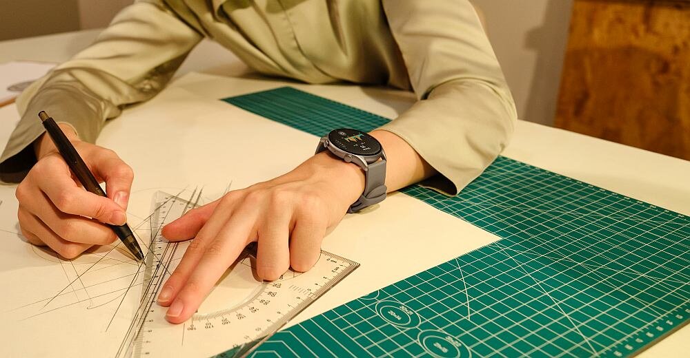 Osoba rysuje linie na papierze za pomocą linijki i ołówka, nosząc smartwatch na nadgarstku. Na stole znajduje się zielona mata do cięcia z białymi liniami pomocniczymi.
