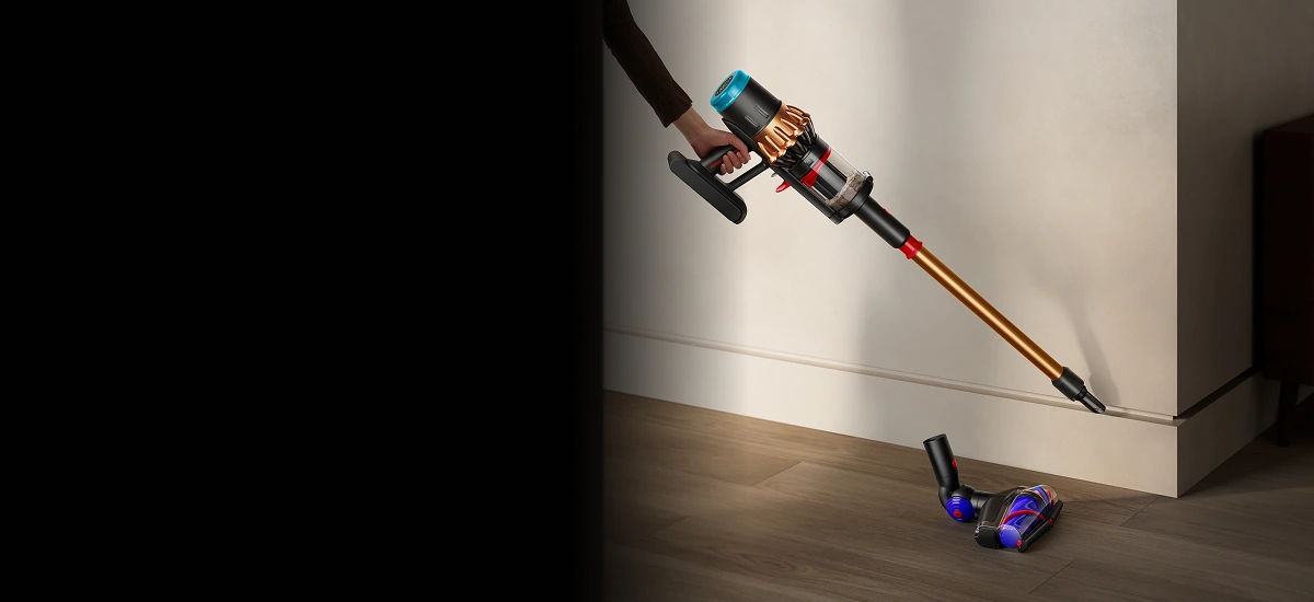 Osoba trzyma odkurzacz o złotej rurze i kolorowym pojemniku, skierowany w stronę podłogi. Na podłodze leżą dwie końcówki odkurzacza.