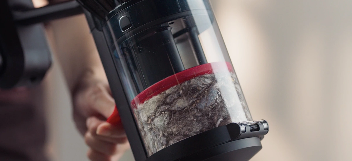 Zbliżenie na przezroczysty pojemnik odkurzacza wypełniony kurzem i włosami. W tle widoczna jest ręka trzymająca urządzenie.