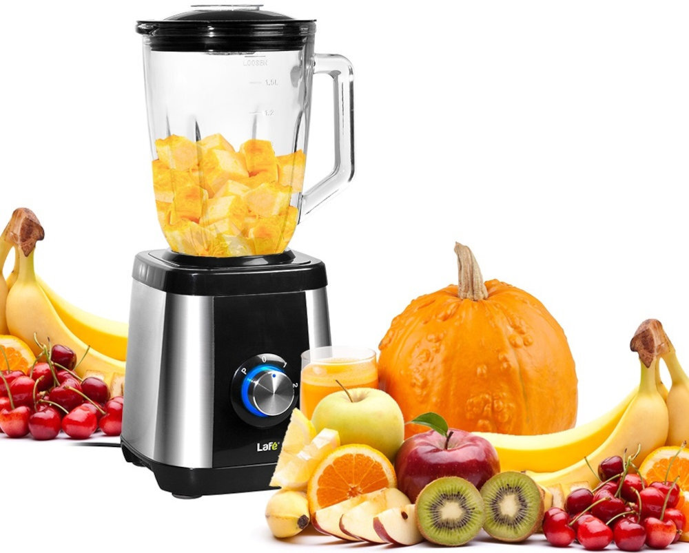 Blender kielichowy w kolorze srebrno-czarnym z pokrojonymi owocami w środku. Wokół blendera znajdują się różne owoce, takie jak banany, pomarańcze, kiwi, jabłka, wiśnie oraz dynia.