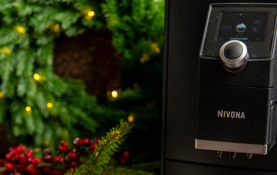 Ekspres do kawy z widocznym logo NIVONA i ekranem z napisem CAPPUCCINO. W tle zielone gałązki z żółtymi światełkami i czerwonymi jagodami.