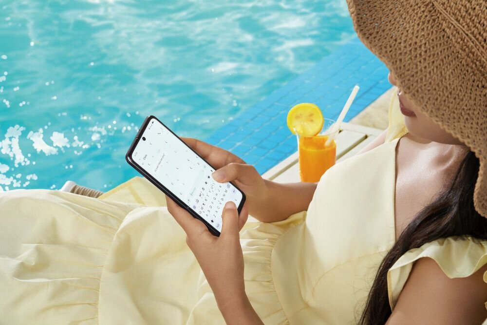 Osoba w kapeluszu siedzi przy basenie, trzymając smartfon z widoczną klawiaturą ekranową. Obok znajduje się szklanka z pomarańczowym napojem i plasterkiem cytryny.