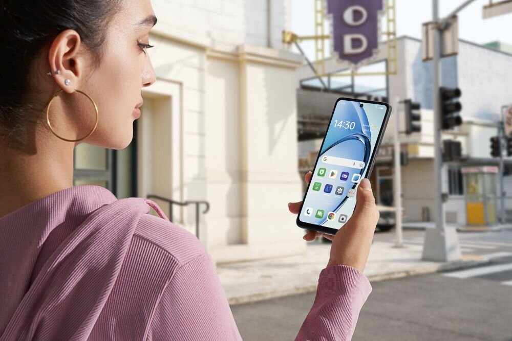 Osoba trzyma smartfon z wyświetlonym ekranem głównym, na którym widać kilka ikon aplikacji i godzinę 14:30. W tle znajduje się ulica z budynkami i sygnalizacją świetlną.