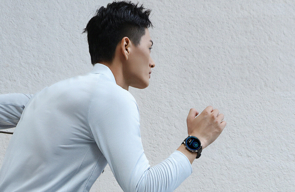 Osoba ubrana w jasną koszulkę biegnie, na nadgarstku ma smartwatch z okrągłym wyświetlaczem. Tło jest jednolite, w jasnym kolorze.