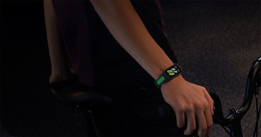 Osoba trzymająca rower ma na nadgarstku czarny smartband z kolorowym wyświetlaczem. Na ekranie widoczne są cyfry i ikony.