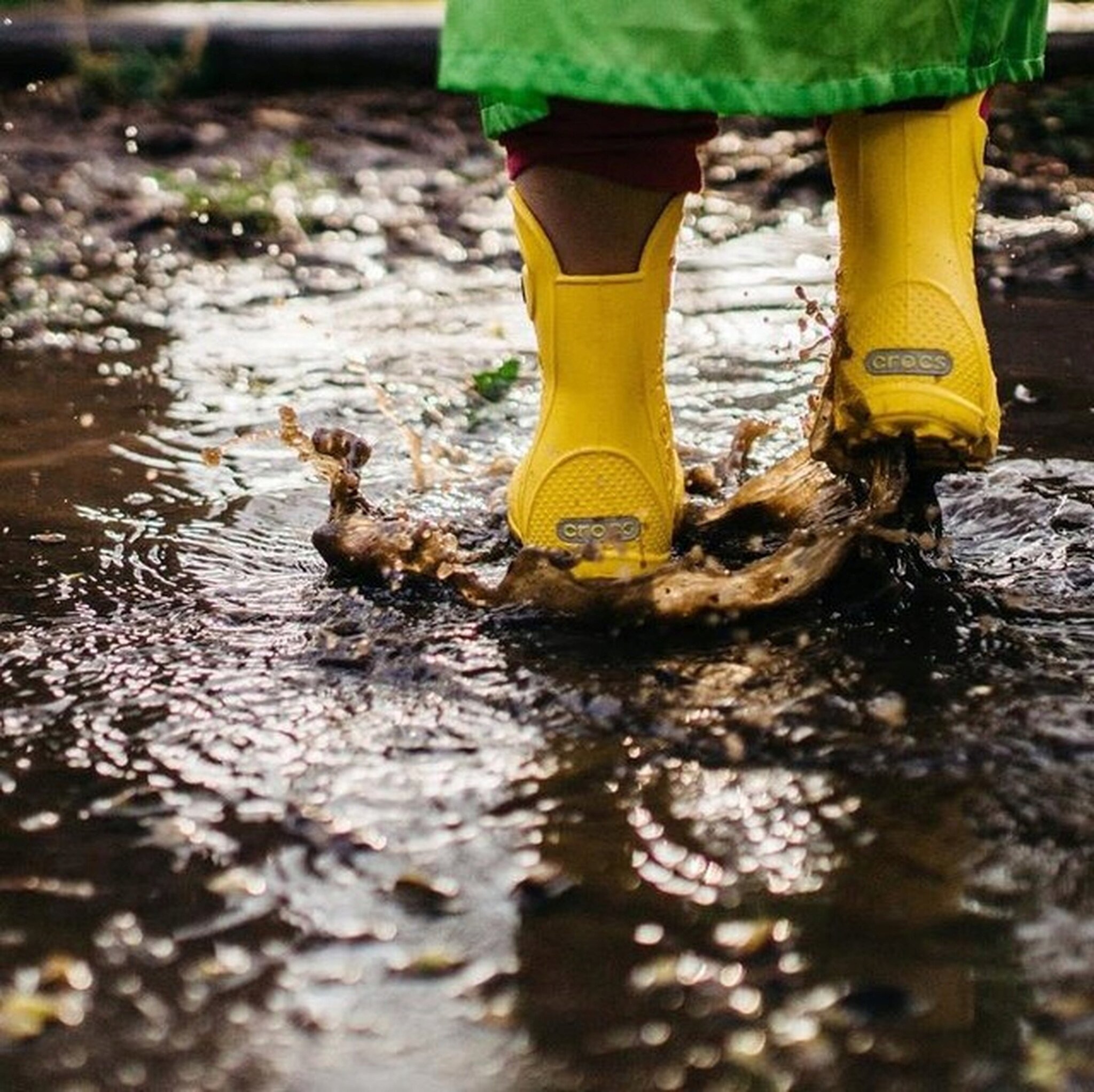 Osoba w żółtych kaloszach skacze po kałuży, rozpryskując wodę. Na butach widoczne jest logo \'crocs\'.