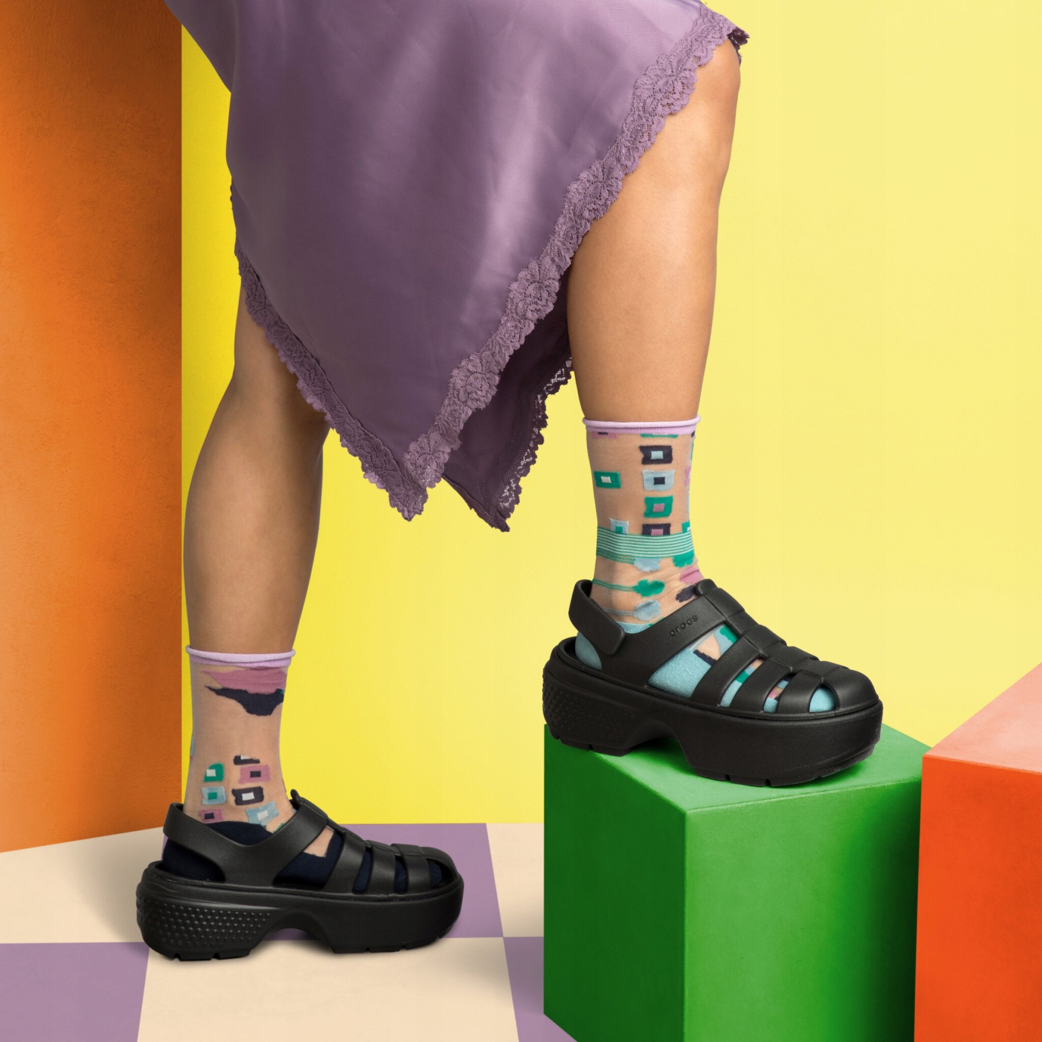 Osoba nosi czarne sandały na platformie z widocznym logo \'Crocs\', stojąc na kolorowych geometrycznych blokach. Ma na sobie kolorowe skarpetki z wzorami oraz fioletową spódnicę z koronkowym wykończeniem.