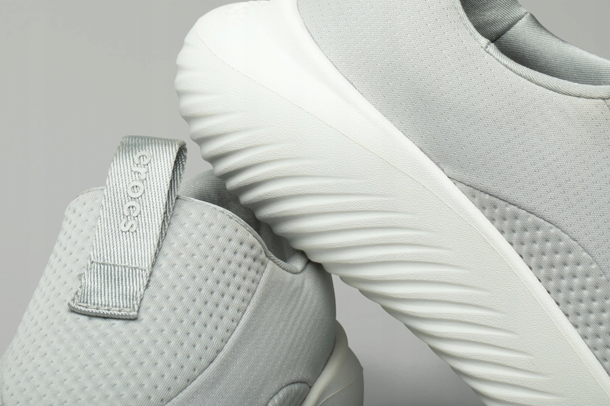 Na obrazie widoczne są jasnoszare buty sportowe z teksturowaną powierzchnią i białą podeszwą. Na jednym z butów znajduje się napis \'crocs\'.