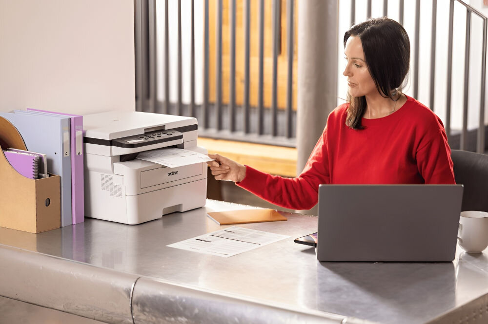 Urządzenie wielofunkcyjne BROTHER DCP-B7600D Automatyczny druk dwustronny prezentacja urządzenia podczas korzystania kobieta wyciągająca kartkę z urządzenia prędkość druku tryb monochromatyczny eliminacja długiego czasu oszekiwania czas potrzebny na wydruk pierwszej strony
