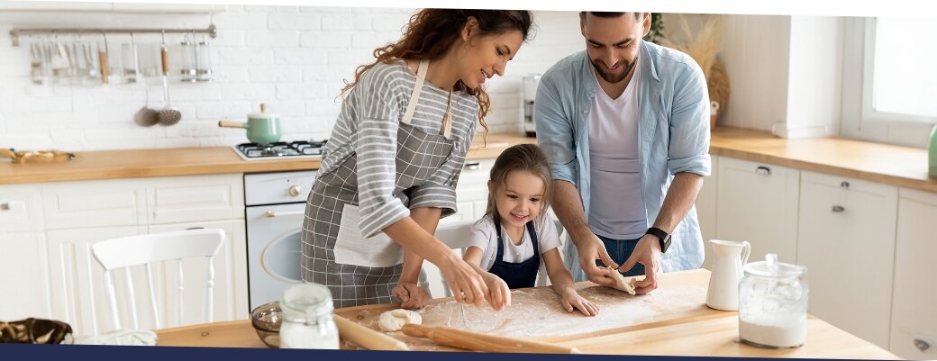 Trzy osoby przygotowują ciasto na drewnianym blacie w jasnej kuchni. Na blacie znajdują się wałek, mąka i dzbanek.