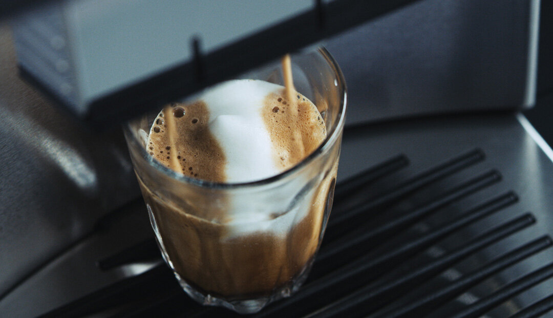 Szklanka z kawą z pianką znajduje się na metalowej kratce ekspresu do kawy. Strumień kawy wlewa się do szklanki z dyszy ekspresu.
