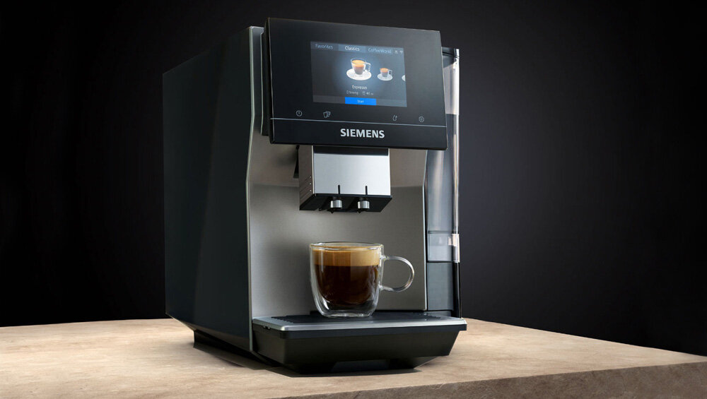 Na obrazie widoczny jest ekspres do kawy z logo SIEMENS, z wyświetlaczem pokazującym dwa rodzaje kawy. Pod dyszami znajduje się szklanka z przygotowaną kawą.