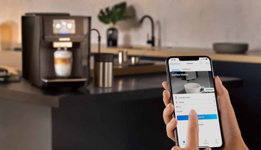 Na blacie kuchennym stoi ekspres do kawy z filiżanką pod dyszą. Osoba trzyma smartfon z aplikacją do sterowania ekspresem, na ekranie widoczny jest tekst: \'Coffee Maker\'.