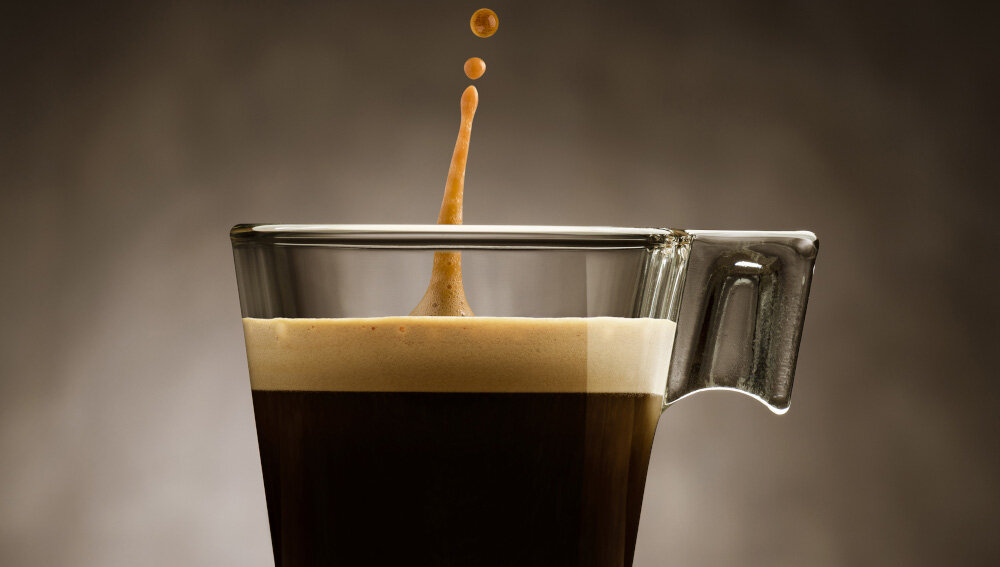 Szklanka z ciemnym napojem, prawdopodobnie kawą, z widoczną pianką na powierzchni. Nad napojem unoszą się krople cieczy.
