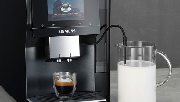 Ekspres do kawy z wyświetlaczem pokazującym filiżankę kawy i tekst \'Your Espresso macchiato is ready!\'. Obok ekspresu stoi dzbanek z mlekiem, a na podstawce znajduje się filiżanka z kawą.