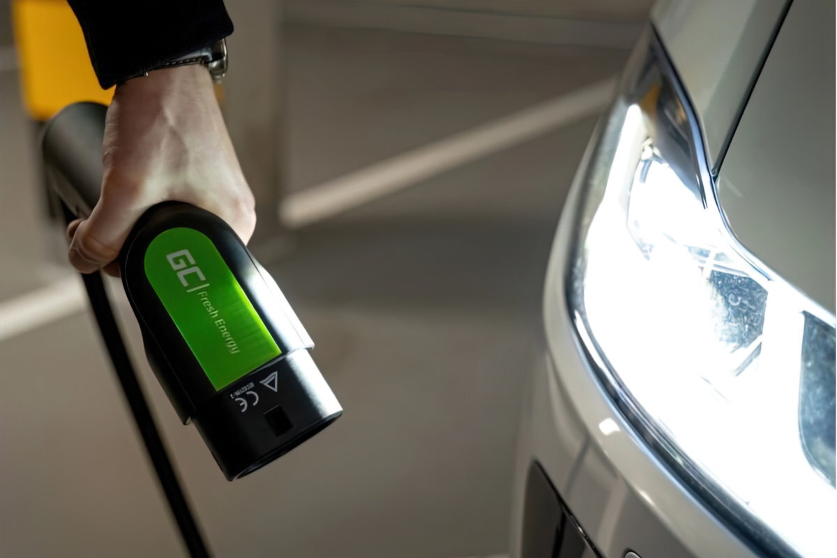 Osoba trzyma wtyczkę kabla do ładowania samochodu elektrycznego z widocznym logo \'GC fresh energy\'. Obok znajduje się reflektor samochodu.