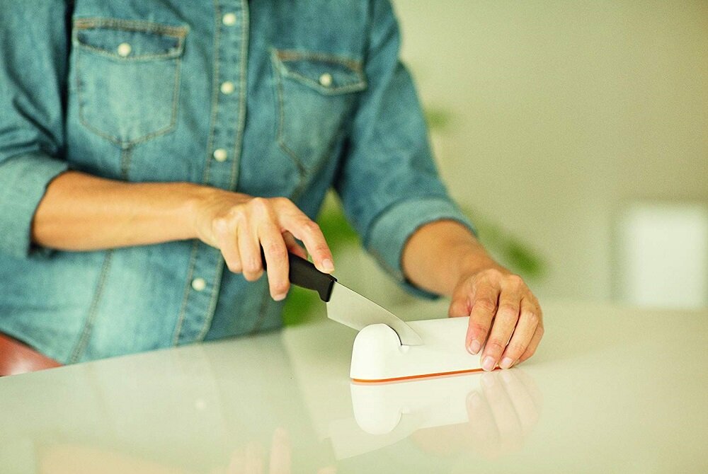 Osoba w dżinsowej koszuli używa białej ostrzałki do noży na gładkiej powierzchni. Ostrzałka ma pomarańczowy spód.