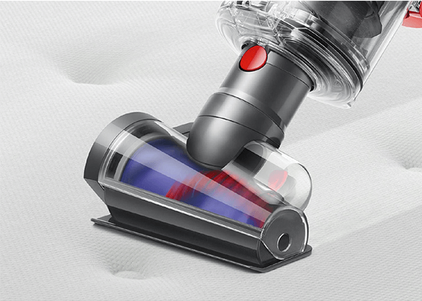 Na obrazie widoczna jest głowica odkurzacza z przezroczystą obudową, w której znajdują się niebieskie i czerwone elementy. Tło stanowi jasna powierzchnia, prawdopodobnie materac.