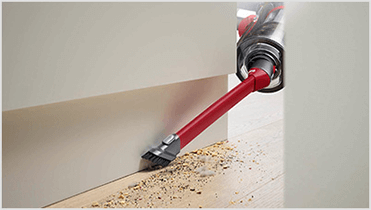 Czerwony odkurzacz z wąską końcówką ssącą usuwa drobinki kurzu i brudu z podłogi przy ścianie.