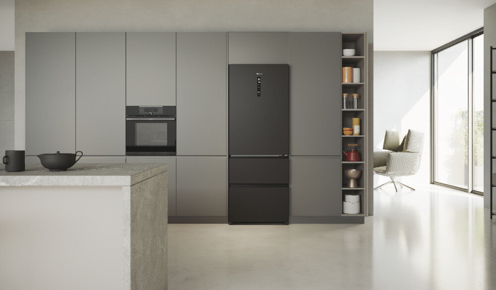 Nowoczesna kuchnia z szarymi szafkami, w której znajduje się czarna lodówka z panelem sterowania na drzwiach. Obok lodówki widoczny jest regał z różnymi przedmiotami, a w tle znajduje się duże okno wpuszczające światło.