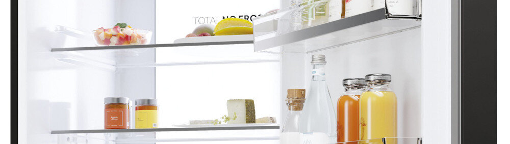 Wnętrze lodówki z widocznymi półkami, na których znajdują się różne produkty spożywcze, takie jak słoiki, butelki z napojami i owoce. Na tylnej ściance widoczny jest napis \'TOTAL NO FROST\'.