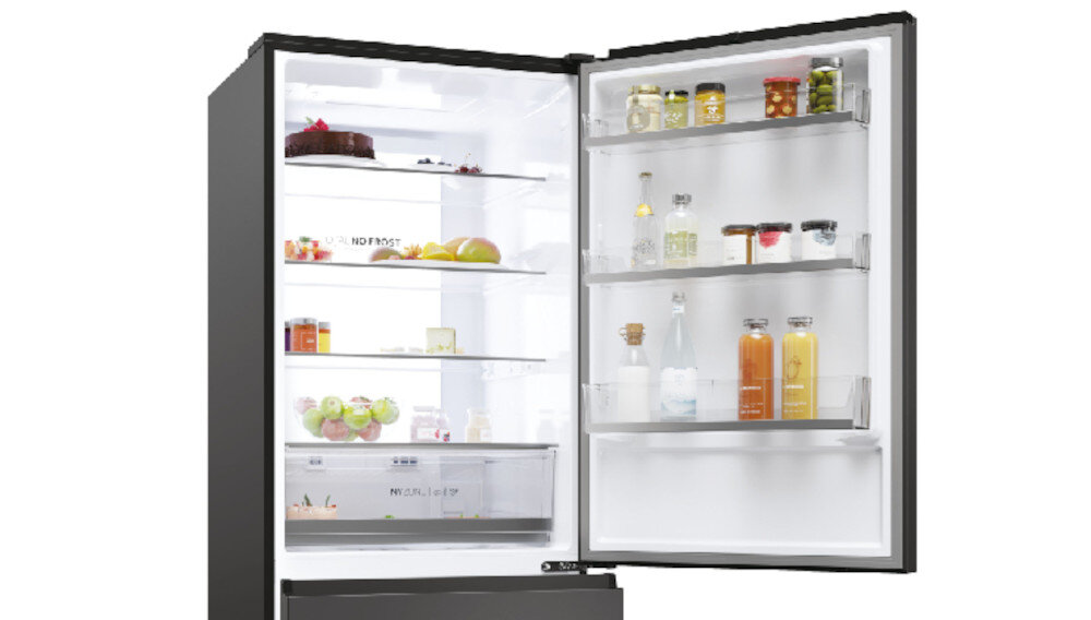 Otwarta lodówka z widocznymi półkami wypełnionymi różnymi produktami spożywczymi, takimi jak owoce, słoiki i butelki. Wnętrze jest jasno oświetlone, a na drzwiach znajdują się dodatkowe półki z napojami i słoikami.