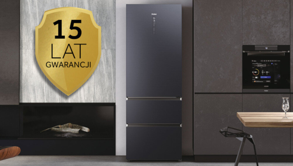 Na obrazie znajduje się czarna lodówka marki Haier z trzema drzwiami, umieszczona w nowoczesnej kuchni. Obok lodówki widoczna jest tarcza z napisem \'15 LAT GWARANCJI\'.
