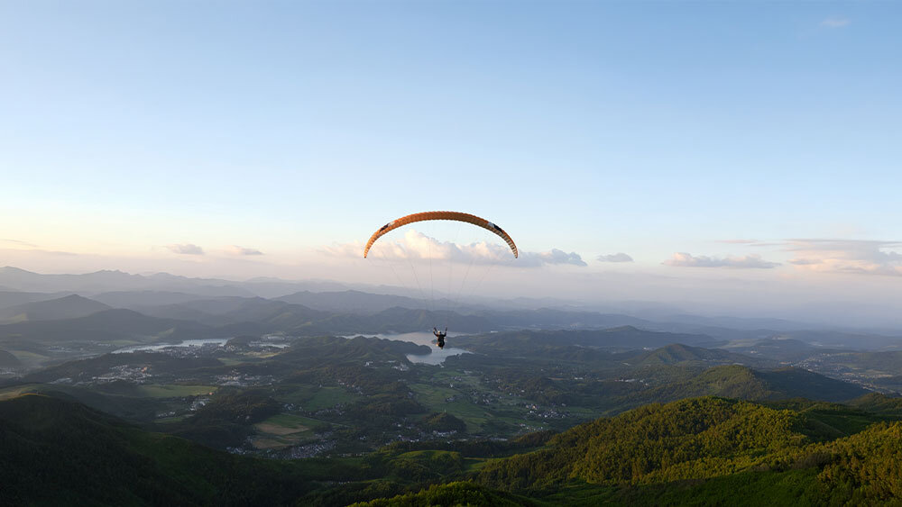 Osoba na paralotni unosi się nad malowniczym krajobrazem z widokiem na góry i doliny oświetlone zachodzącym słońcem.