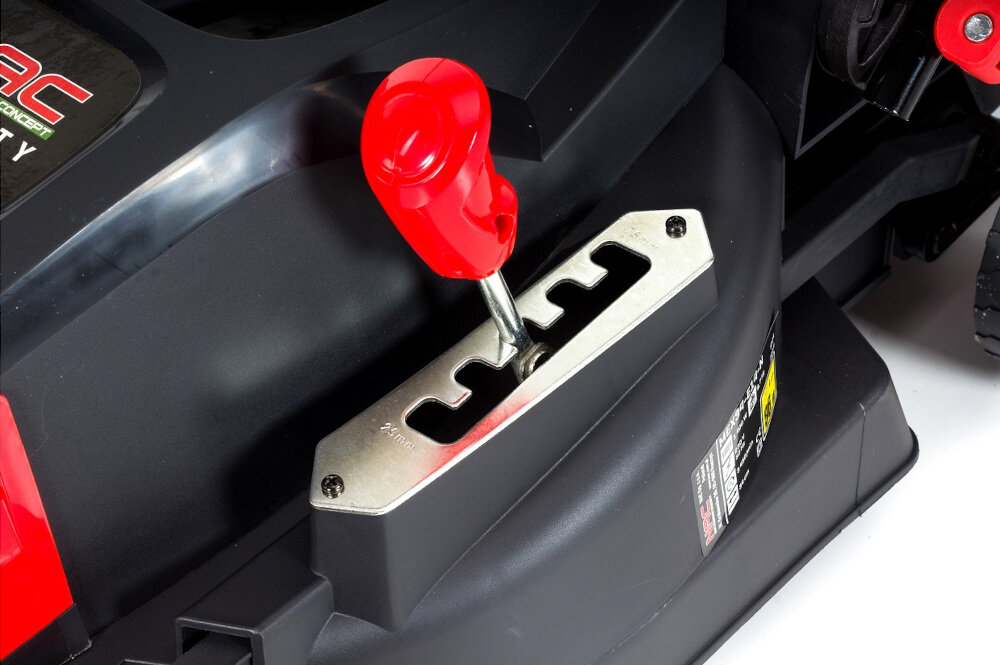 Zbliżenie na część kosiarki elektrycznej z czerwonym uchwytem regulacyjnym i metalową płytką z wycięciami. Widoczne jest logo \'NAC\' na czarnym tle.