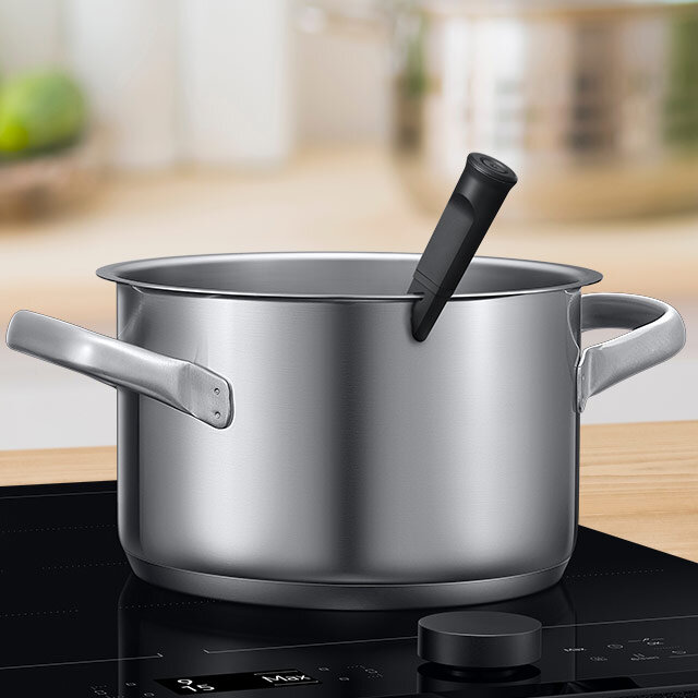 Na obrazie widoczny jest metalowy garnek z uchwytami, stojący na płycie indukcyjnej. W garnku znajduje się czarna termosonda.