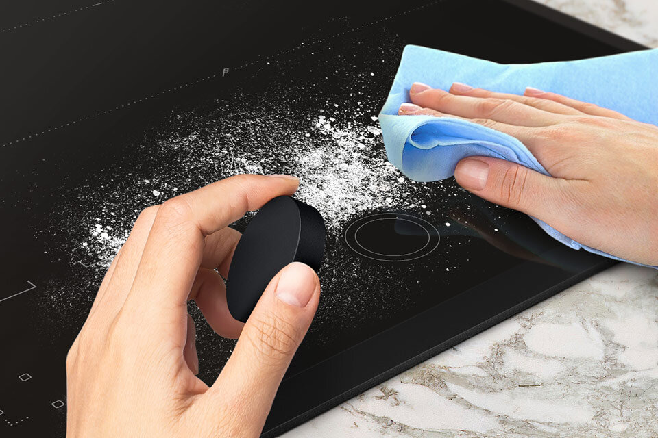 Na obrazie widać czarną płytę indukcyjną, na której jedna ręka trzyma okrągły czarny przedmiot, a druga ręka wyciera powierzchnię niebieską ściereczką. Na płycie znajduje się biały proszek, który jest usuwany.