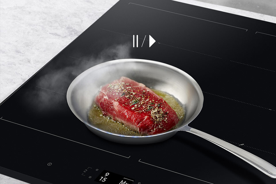 Na płycie indukcyjnej znajduje się patelnia z kawałkiem mięsa, które jest przyprawione i smażone. Na płycie widoczny jest symbol pauzy i odtwarzania oraz cyfry \'9\' i \'15\'.
