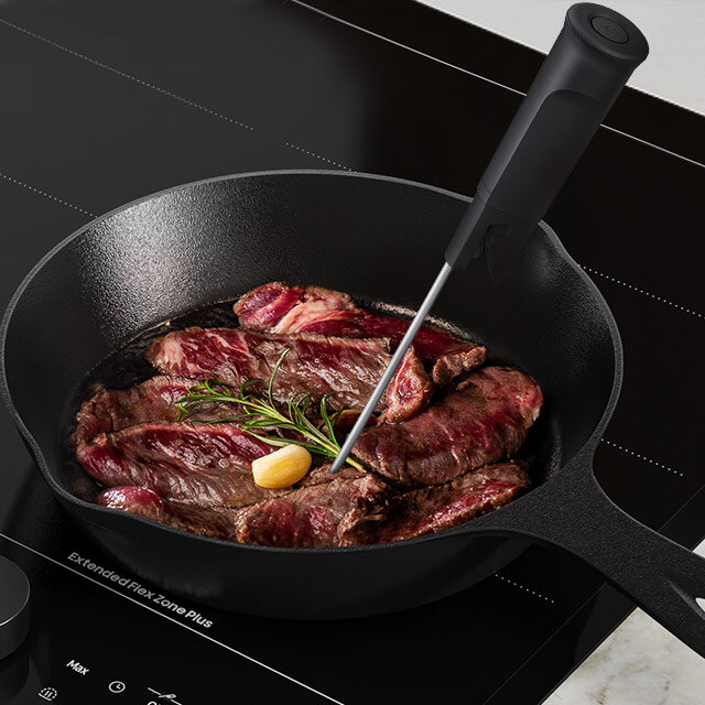Na płycie indukcyjnej znajduje się patelnia z kawałkami mięsa, ziołami i czosnkiem, a w mięso wbita jest termosonda. Na płycie widoczny jest napis \'Extended Flex Zone Plus\'.
