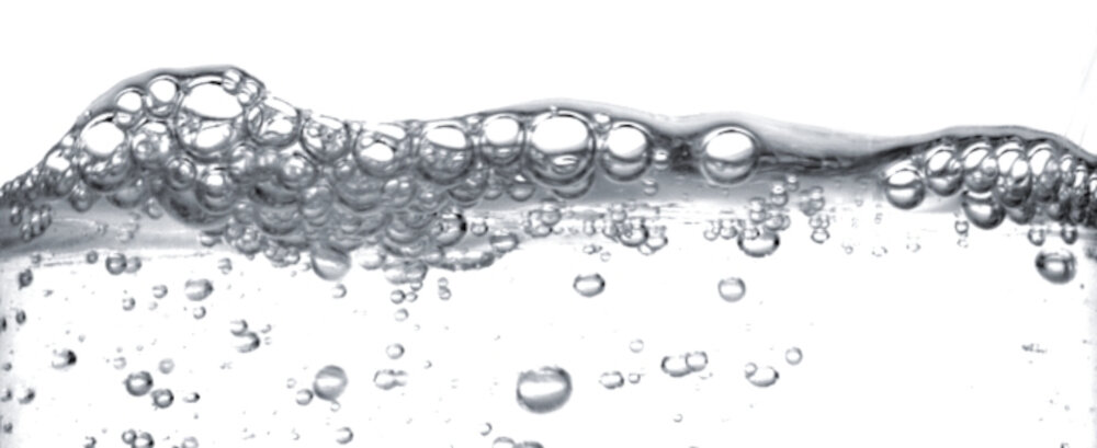 Obraz przedstawia przezroczystą ciecz z licznymi bąbelkami unoszącymi się na powierzchni. Tło jest białe, co podkreśla efekt bąbelków.