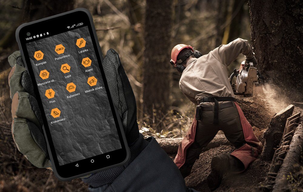 Osoba w kasku i ochronnym ubraniu używa piły łańcuchowej w lesie. Na pierwszym planie widoczny jest smartfon z aplikacjami takimi jak Kompas, Poziomica, Latarka, Wysokościomierz, Lupa, Alarm, Pion, Kalkulator, Miernik dźwięku, Krokomierz.