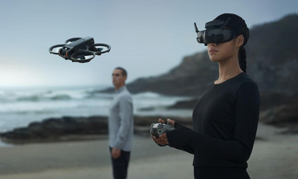Osoba w okularach VR trzyma kontroler, a w powietrzu unosi się dron. W tle widać plażę i morze.