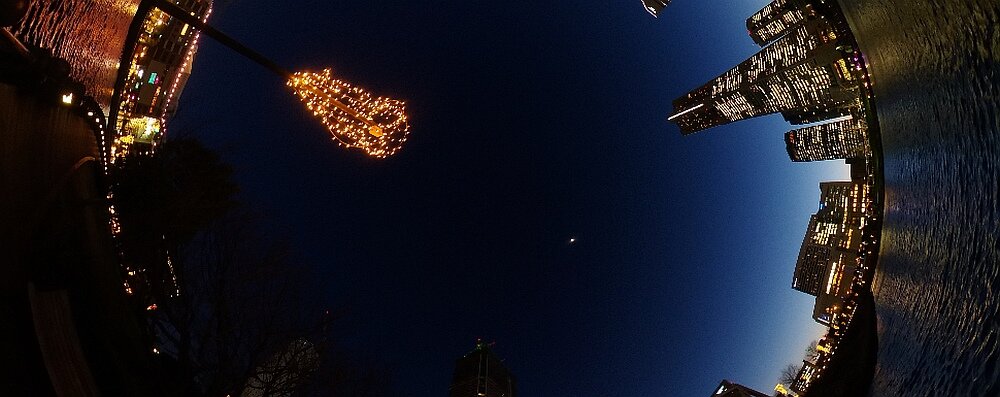 Panorama miasta nocą z widocznymi oświetlonymi budynkami i lampami ulicznymi. Na niebie widać księżyc.