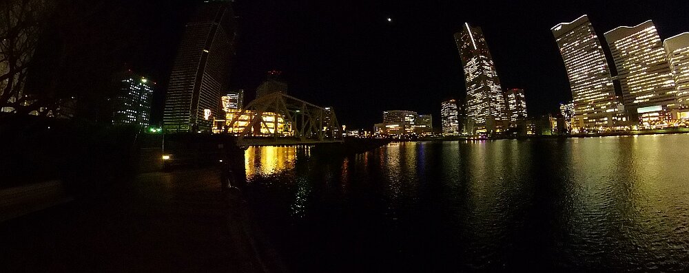 Nocna panorama miasta z oświetlonymi wieżowcami odbijającymi się w wodzie. Na niebie widoczny jest księżyc.