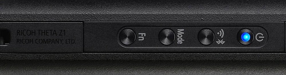 Zbliżenie na panel boczny kamery z przyciskami oznaczonymi jako Fn, Mode, ikoną Bluetooth oraz przyciskiem z niebieskim podświetleniem. Widoczny jest tekst: \'RICOH THETA Z1 RICOH COMPANY, LTD.\'.