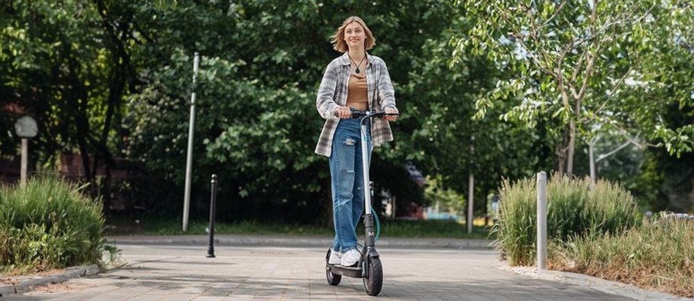 Osoba jedzie na szarej hulajnodze elektrycznej po wybrukowanej ścieżce, w tle widać drzewa i krzewy.
