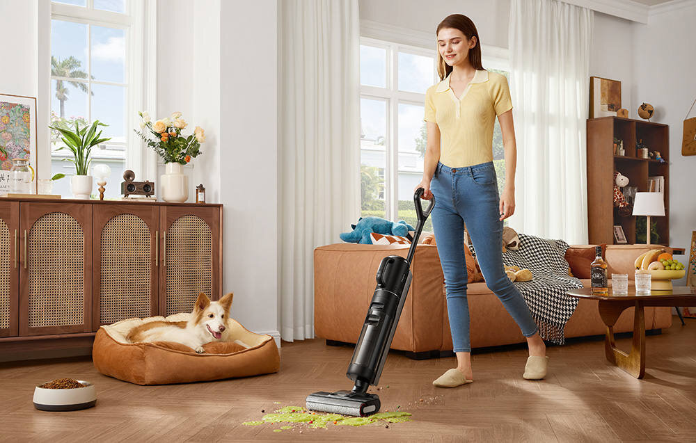 Osoba używa odkurzacza mopującego na drewnianej podłodze, obok leży pies na legowisku. W tle znajduje się sofa z poduszkami i stolik z owocami oraz książkami.