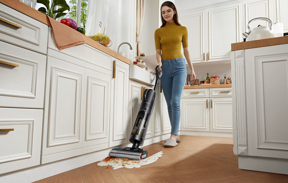 Kobieta używa odkurzacza mopującego na drewnianej podłodze w kuchni, czyszcząc rozlany płyn z resztkami jedzenia. W tle widać jasne szafki kuchenne i zlew z rośliną na blacie.