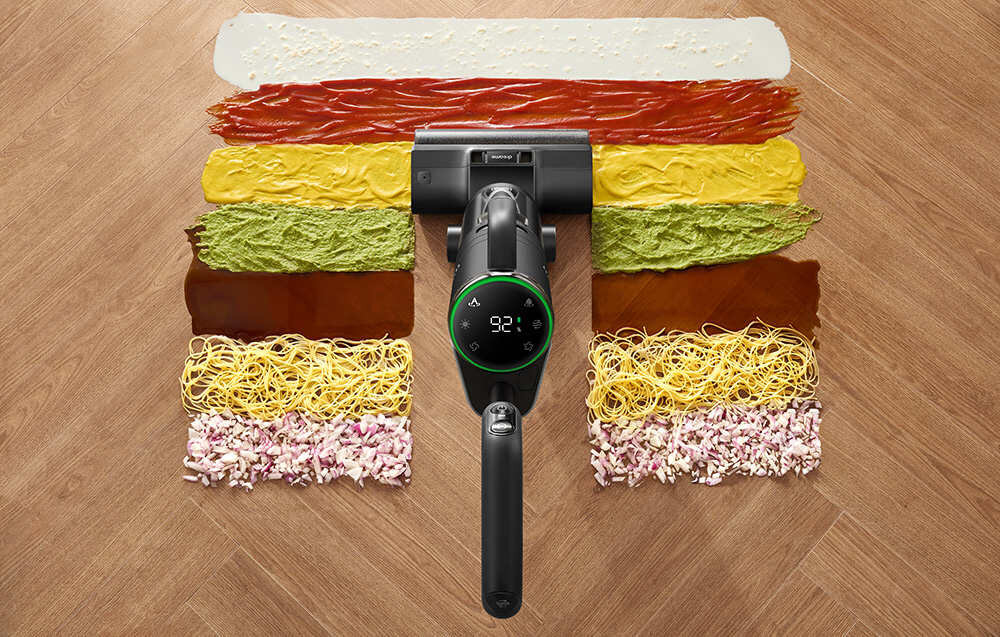 Odkurzacz mopujący z wyświetlaczem pokazującym \'92%\' znajduje się na drewnianej podłodze, czyszcząc różne rodzaje zabrudzeń ułożone w pasy, takie jak sosy, warzywa i makaron.