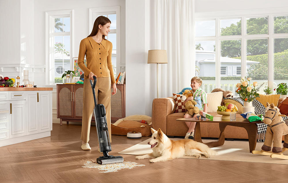 Kobieta używa odkurzacza mopującego na drewnianej podłodze, obok leży pies. W tle widać dziecko siedzące na kanapie z zabawkami i kwiatami na stole.