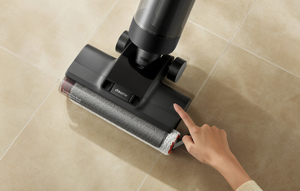 Odkurzacz mopujący z widocznym logo \'dreame\' na głowicy, używany na jasnej podłodze. Osoba wskazuje palcem na część urządzenia.