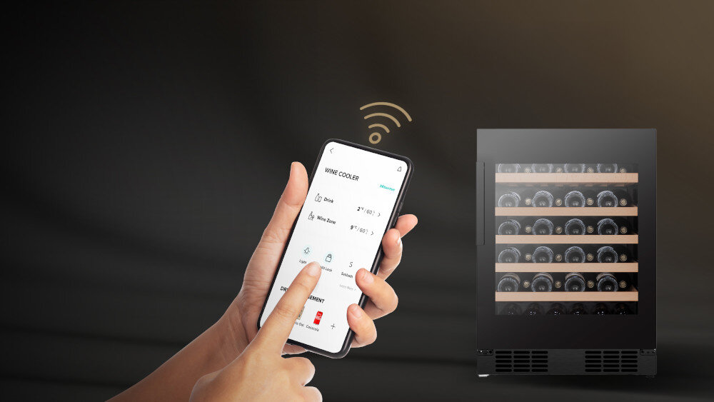Osoba trzyma smartfon z aplikacją do sterowania chłodziarką do wina, na ekranie widoczny jest tekst \'WINE COOLER\'. Obok znajduje się chłodziarka z kilkoma półkami wypełnionymi butelkami wina.