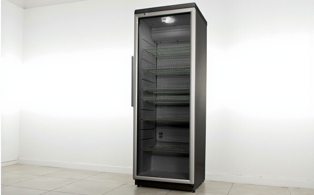 Witryna chłodnicza o metalicznym wykończeniu z przeszklonymi drzwiami i kilkoma półkami w środku. Stoi w jasnym pomieszczeniu na płytkach podłogowych.