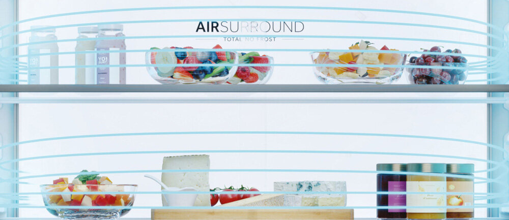 Wnętrze lodówki z półkami wypełnionymi różnorodnymi produktami spożywczymi, takimi jak owoce, sery i napoje. Na górnej półce widoczny jest napis \'AIR SURROUND TOTAL NO FROST\'.