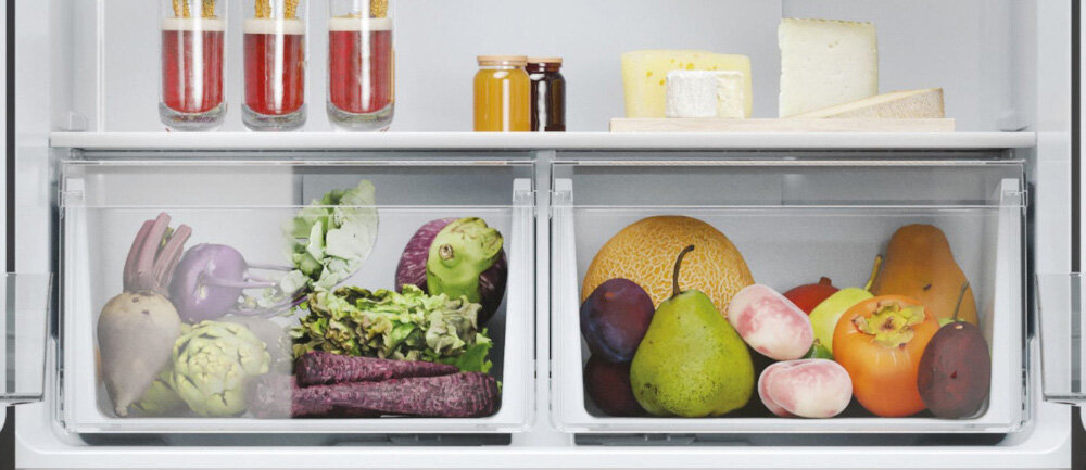 Wnętrze lodówki z dwoma przezroczystymi szufladami wypełnionymi różnorodnymi warzywami i owocami. Na górnej półce znajdują się słoiki z czerwonym płynem, małe butelki oraz kawałki sera.