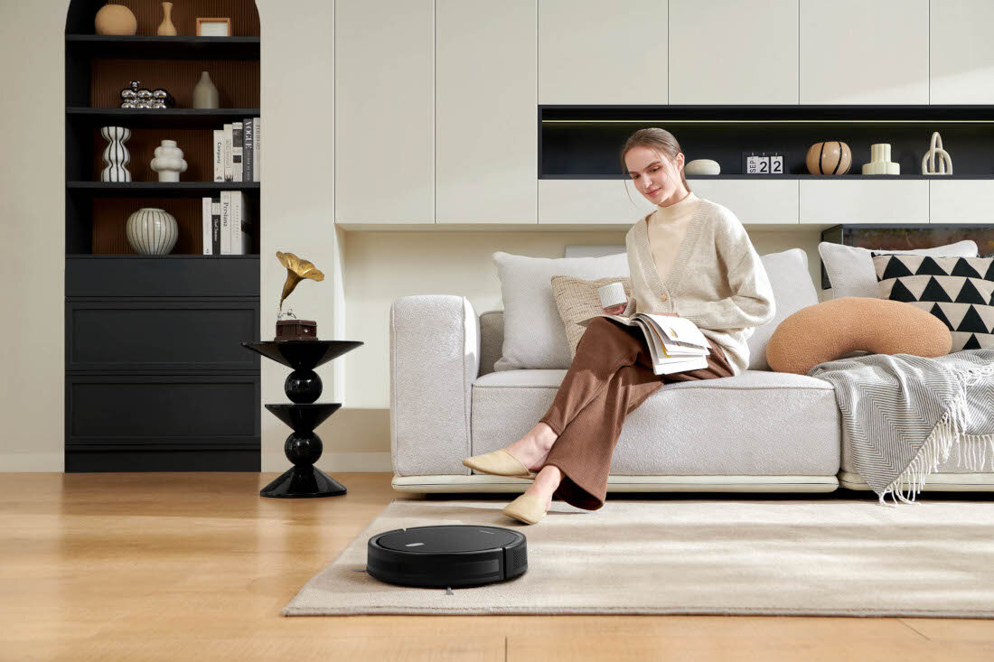 W jasnym salonie na dywanie znajduje się czarny robot sprzątający. Na kanapie siedzi osoba z książką, a w tle widać półki z dekoracjami.